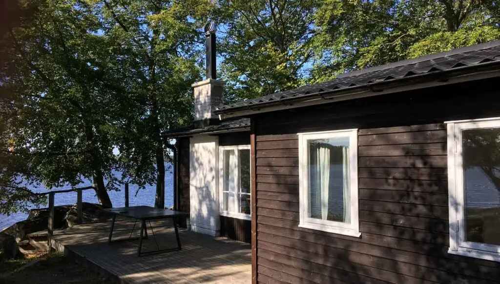 Stuga vid sjön i Åsnens Nationalpark med träterrass, skog och lugnt vatten i bakgrunden en solig dag.