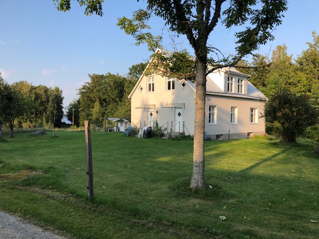 Ljust hus med klassisk fasad omgivet av gräsmatta, träd och lantlig natur i lugn miljö.