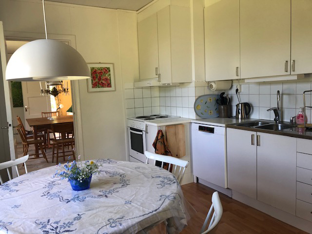 Ljust kök med vita skåpluckor, matbord för flera personer, spis, diskmaskin och arbetsytor i hemmiljö.