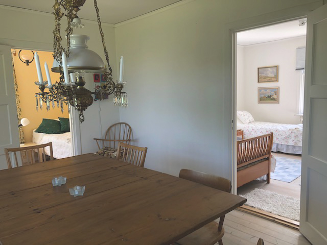 Matsal med stort träbord, klassisk taklampa och öppning mot sovrum i ljust och hemtrevligt hus.