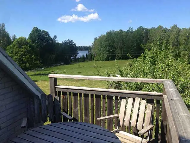Balkong med träbord och stol, utsikt över grön äng, skog och glimt av sjö i lugn, privat miljö.