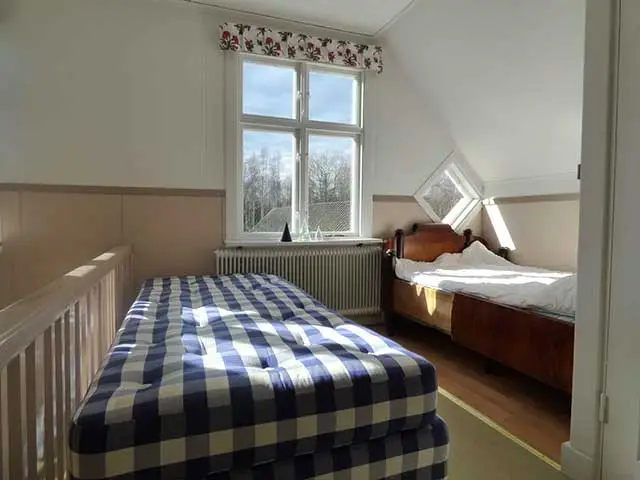 Sovrum med två enkelsängar, snedtak och stort fönster som släpper in rikligt med dagsljus i lugn stugmiljö.