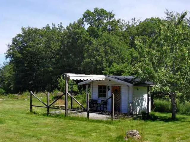 Liten vit stuga i grönskande trädgård med uteplats, omgiven av träd och öppna gräsytor i Småland.
